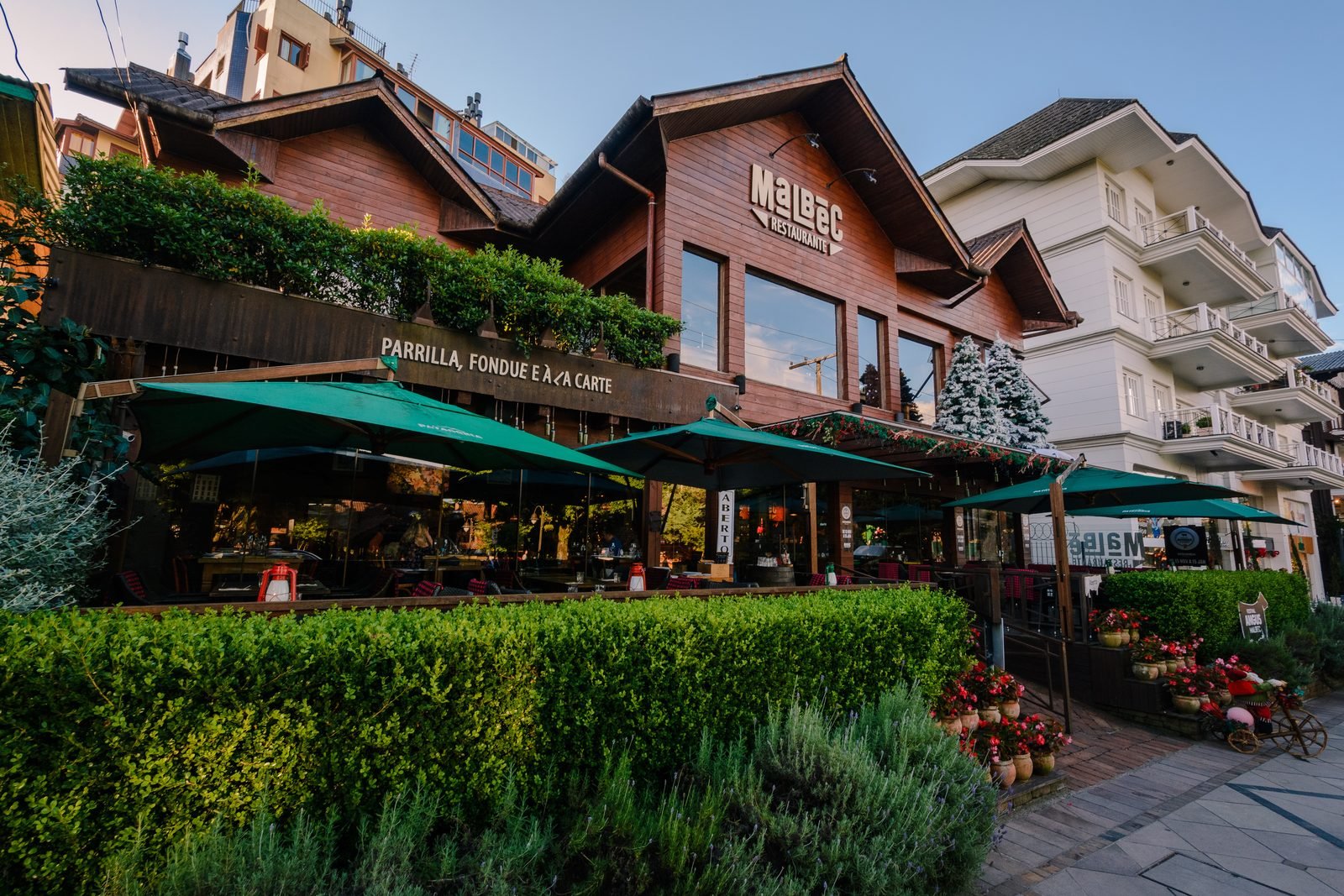 Fachada de chalé de madeira de um restaurante em Gramado ao entardecer, com guarda-sóis verdes sobre a varanda e canteiros floridos na entrada.