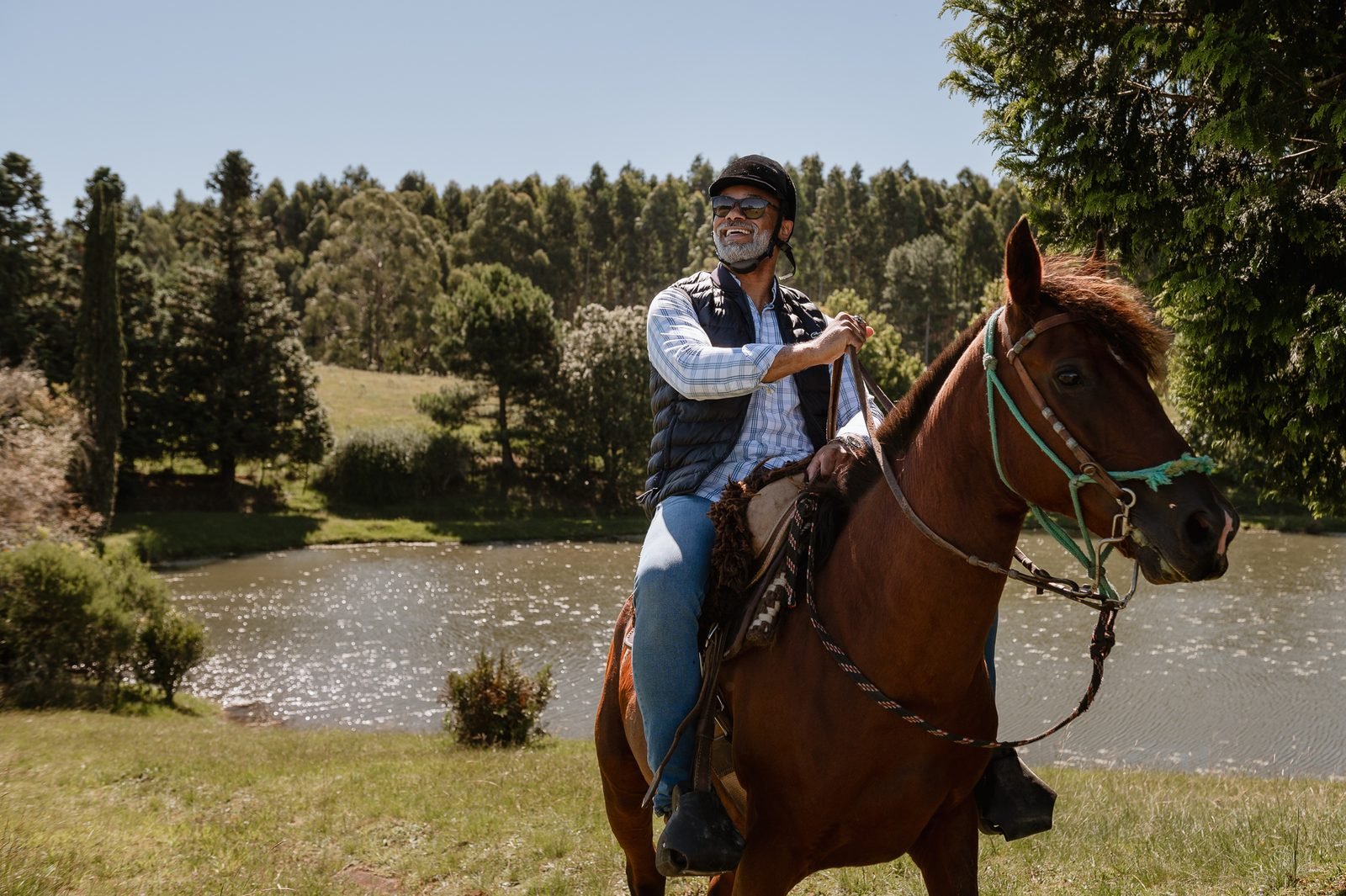 Mulher de capacete de montaria em cavalo, com árvores e açude ao fundo