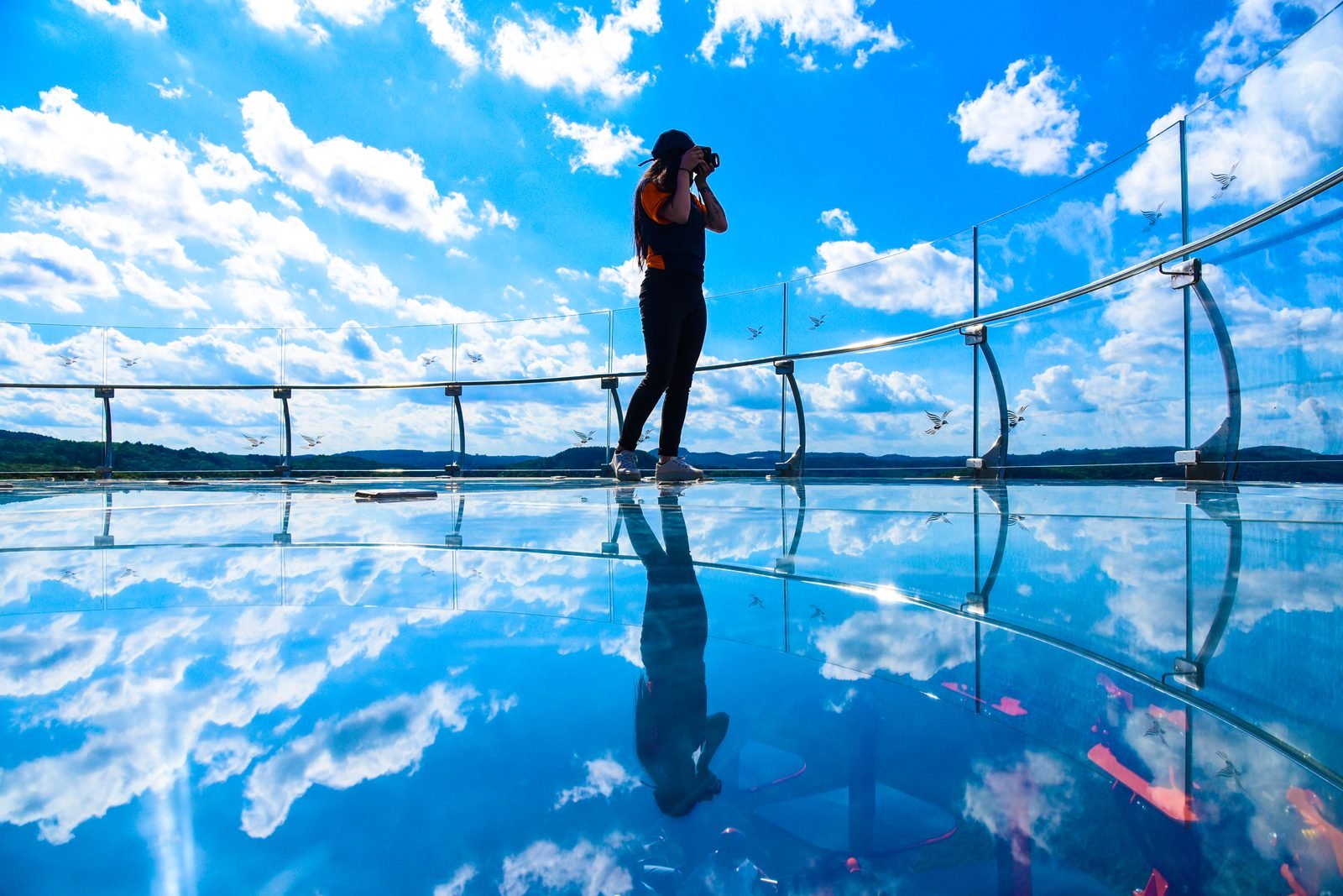 Pessoa de pé sobre o piso de vidro da plataforma fotografando, com o céu e as nuvens refletidos no chão