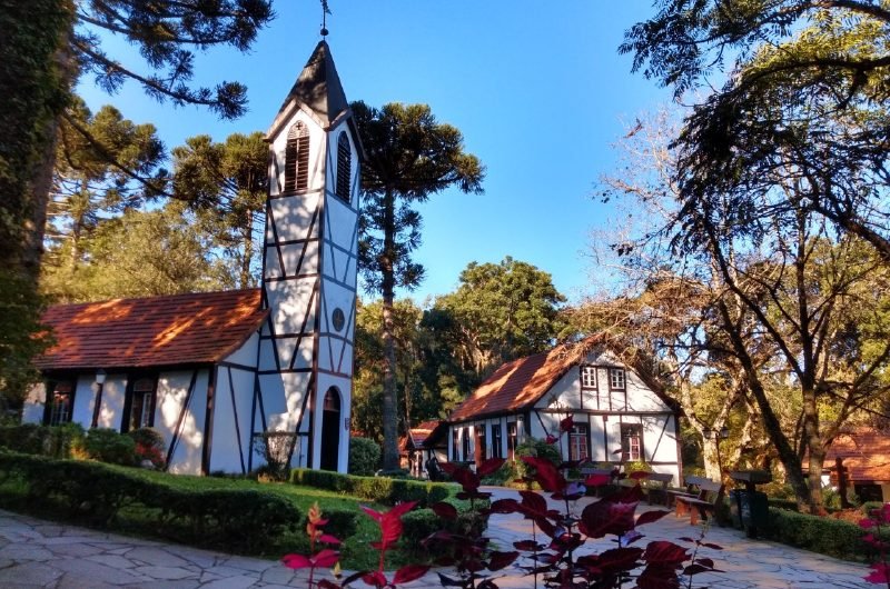 Capela em estilo enxaimel com torre e telhado vermelho, entre araucárias, no Parque Aldeia do Imigrante