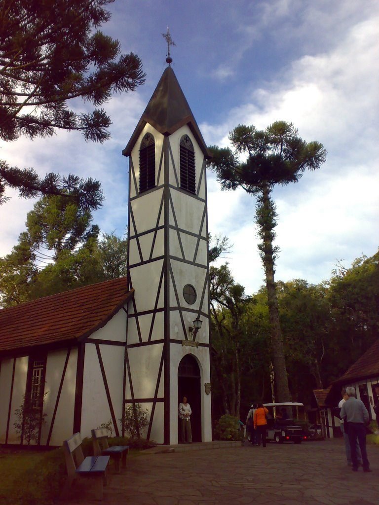 Torre da capela em estilo enxaimel vista de baixo, com araucárias ao fundo e visitantes no caminho de pedra